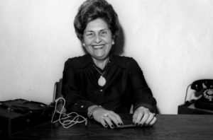 Foto em preto e branco de Dorina de Gouvêa Nowill. Ela está sorrindo, sentada por trás de uma mesa de madeira, com as mãos apoiadas sobre o móvel. Ao fundo, uma parede clara. Dorina é uma mulher branca de cabelos curtos. Ela usa roupa escura, brincos de pérola, colar, pulseira e anéis. Em cima da mesa, do lado direito, há um telefone do modelo antigo de discar, e do lado esquerdo, uma bolsa.